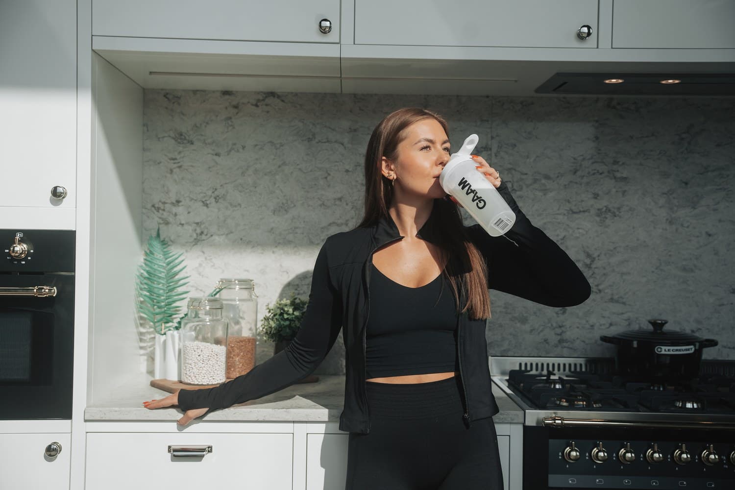 A woman in black training wear drinking from a white GAAM shaker bottle in a bright kitchen